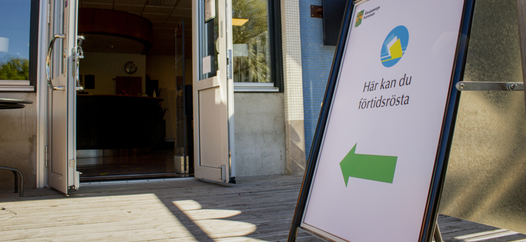 Gatupratare med texten "Här kan du rösta", utanför en lokal med öppen dörr.
